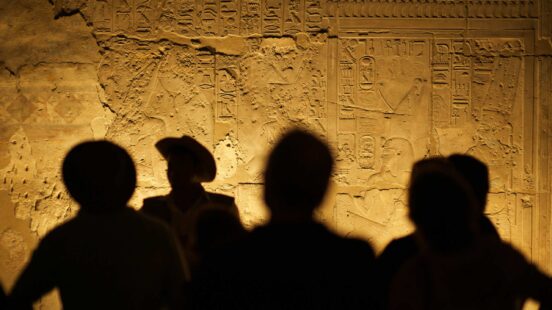 Silhouettes of people viewing an ancient Egyptian wall carving with many rows of hieroglyphs.