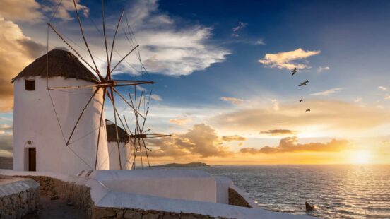 Two traditional white windmills on a coast during a golden sunset with birds flying in the sky.