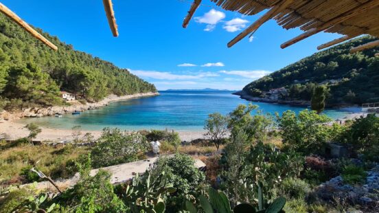 Scenic view of a turquoise bay and pebble beach framed by a rustic wooden shelter and green mountains.