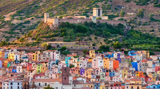 Colorful houses in a coastal village below a historic stone castle on a green hillside.