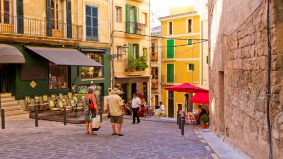 A narrow European street with yellow and green buildings, a sidewalk cafe, and pedestrians walking on cobblestones.
