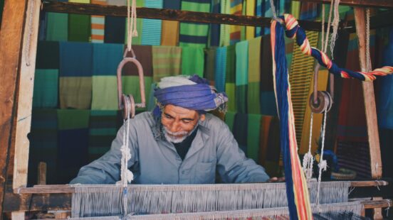 A man working at a traditional wooden loom with rows of colorful green and blue fabrics in the background.
