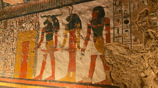 Ancient Egyptian wall art showing three figures with animal heads and rows of hieroglyphics on a textured surface.
