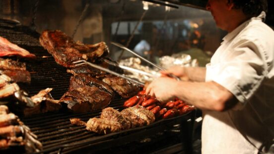 A chef cooks thick cuts of meat and red sausages on a large, glowing Argentinian parrilla or barbecue grill.