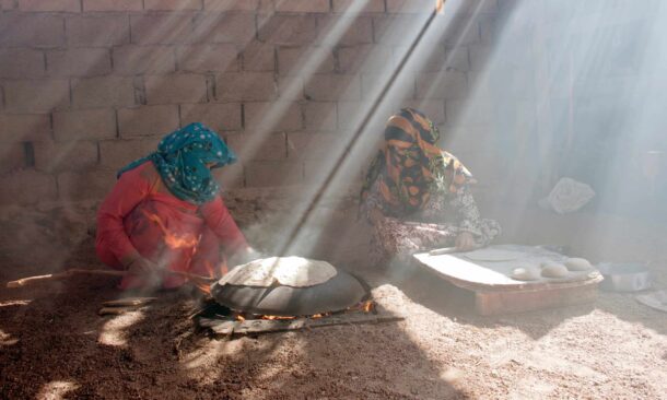 Two people cooking flatbread on a large domed griddle over a fire in a dusty, sunlit courtyard.