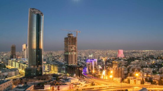 An aerial view of a city at night with illuminated skyscrapers, busy roads, and a clear evening sky.