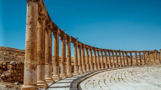 A long perspective of ancient stone columns forming a curve along a paved plaza under a bright blue sky.
