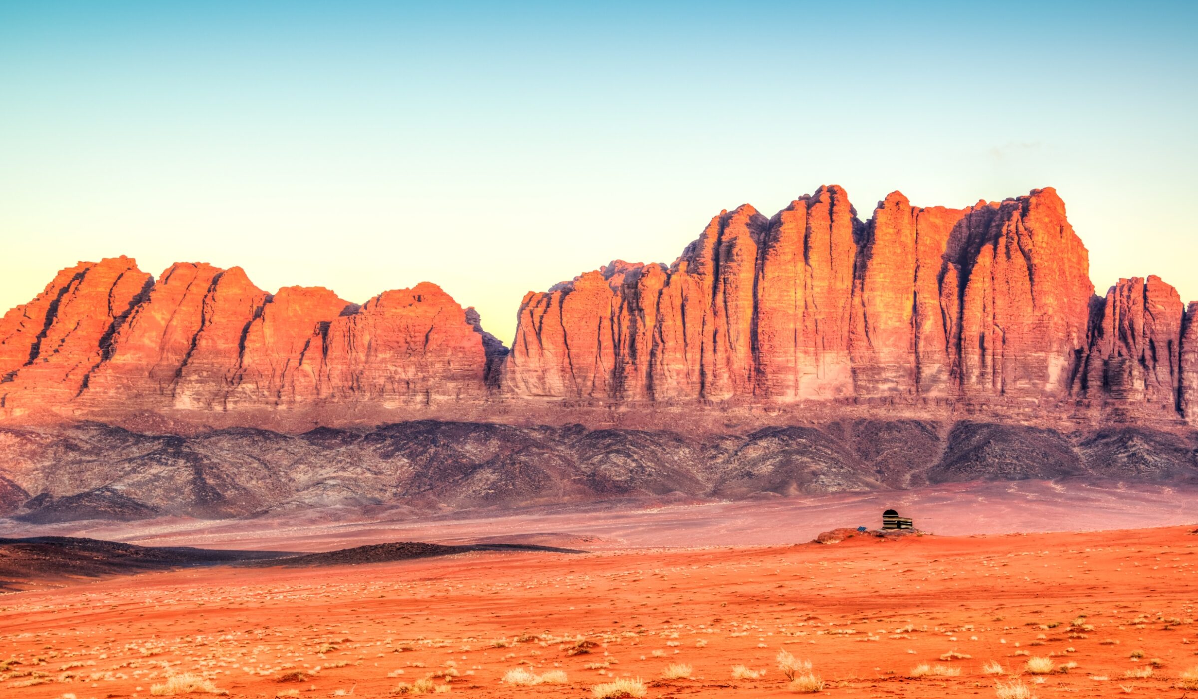 A wide shot of dramatic red rock mountains rising from a flat desert floor at sunset.
