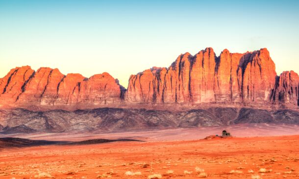 A wide shot of dramatic red rock mountains rising from a flat desert floor at sunset.