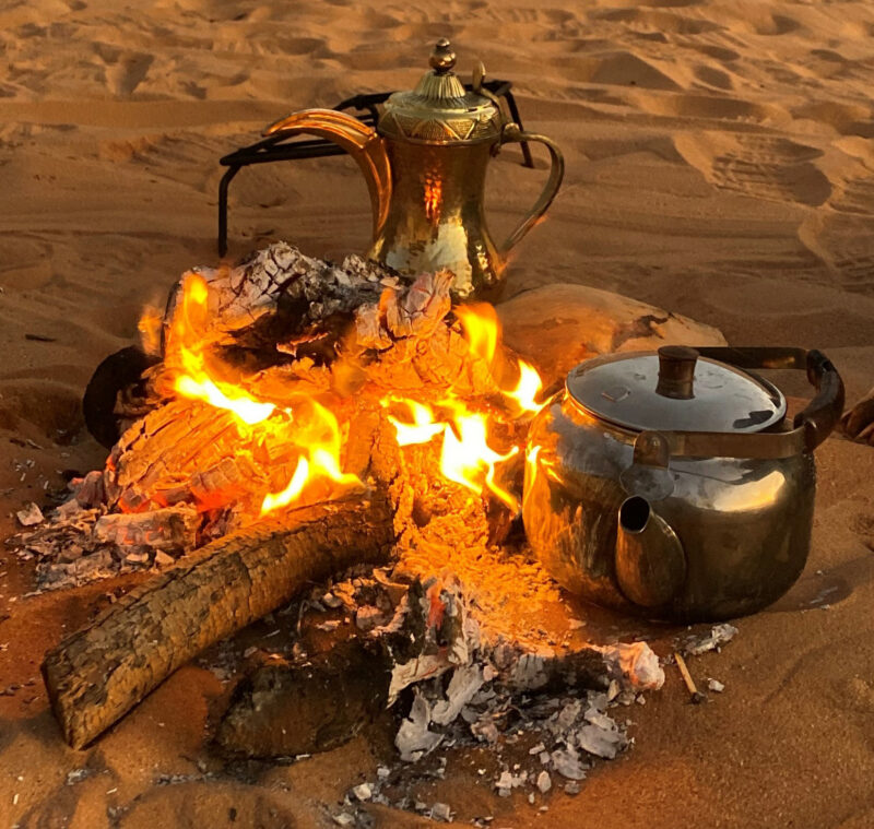 Two brass kettles, a traditional coffee pot and a round tea pot, sit beside a small campfire on the desert sand.