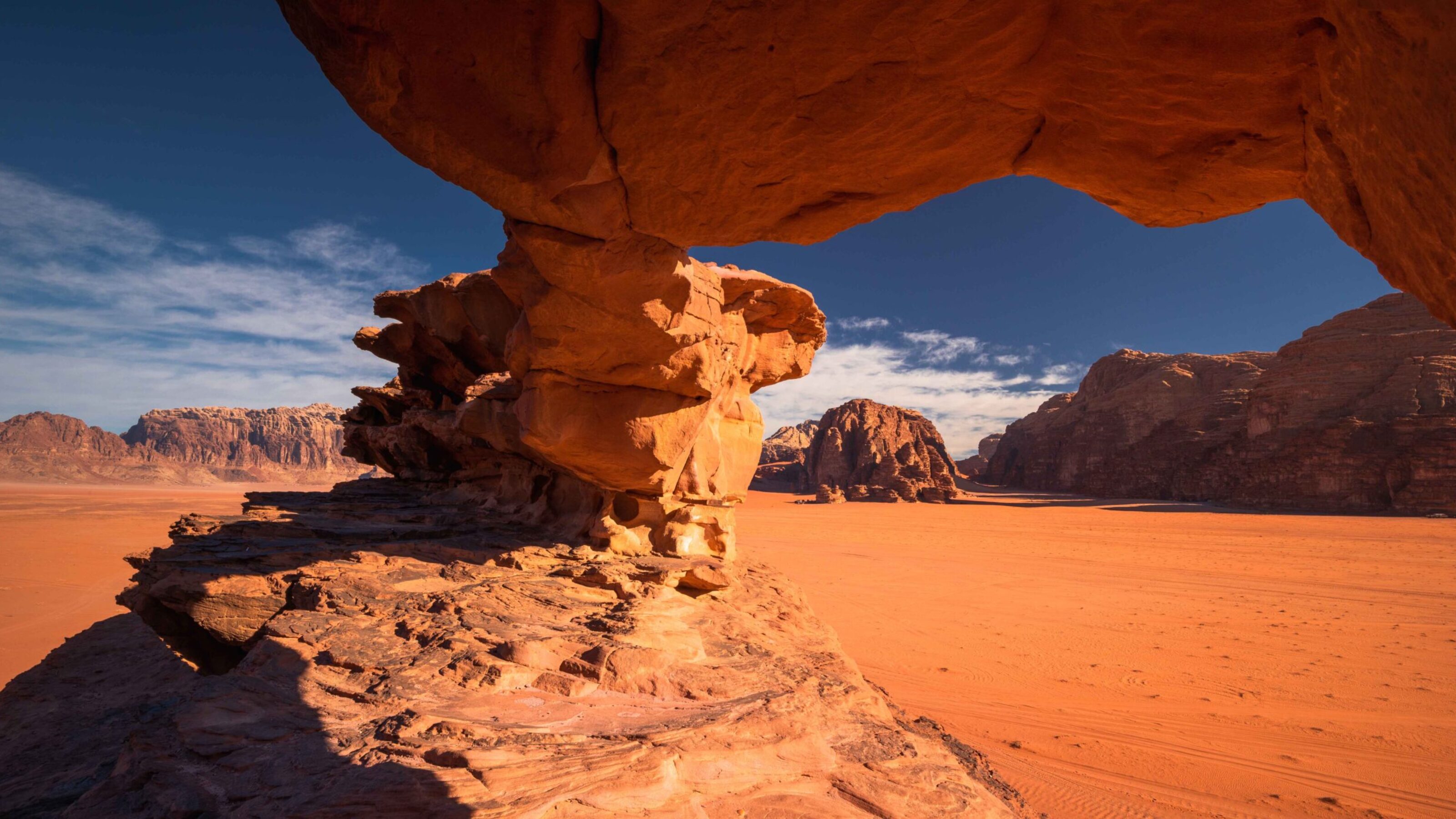 Natural sandstone rock arch formation frames the reddish-orange desert and mountains of Wadi Rum under a deep blue sky.