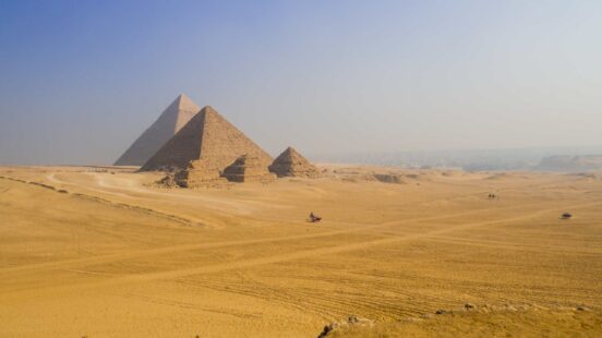 The Great Pyramids of Giza standing in a vast desert landscape under a pale blue sky.