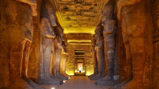 A row of monumental ancient Egyptian statues standing in a dimly lit temple corridor with carved ceilings.