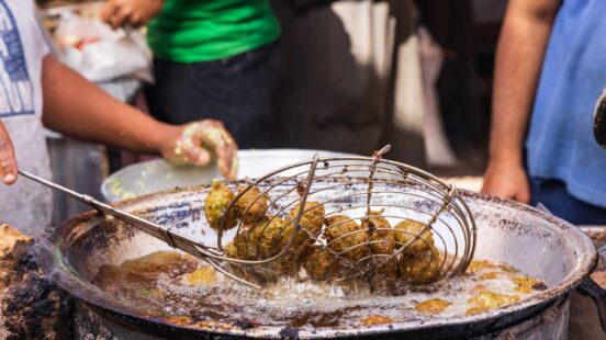 A metal strainer lifting freshly fried falafel from a large pan of bubbling oil in a street food setting.