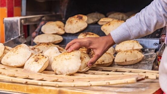 A hand reaching for warm, puffed pita bread arranged on a wooden lattice rack in an outdoor market setting.