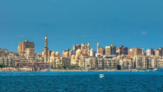 A wide view of a coastal city skyline with mosque domes and minarets against a clear blue sky and ocean.