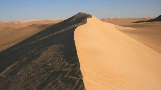 A steep sand dune ridge showing a stark divide between light and shadow under a clear sky in the desert.