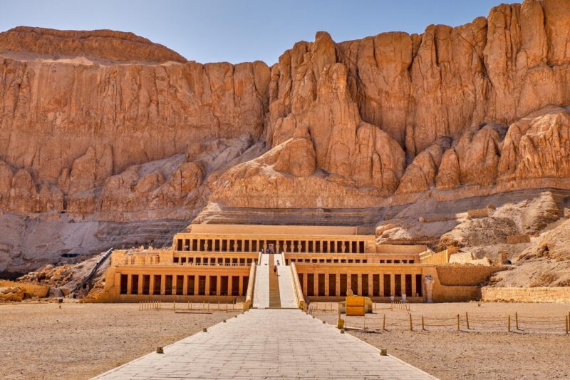 The massive, multi-tiered Mortuary Temple of Hatshepsut built into a sheer cliff face under a bright sky. Luxury Middle East holidays.