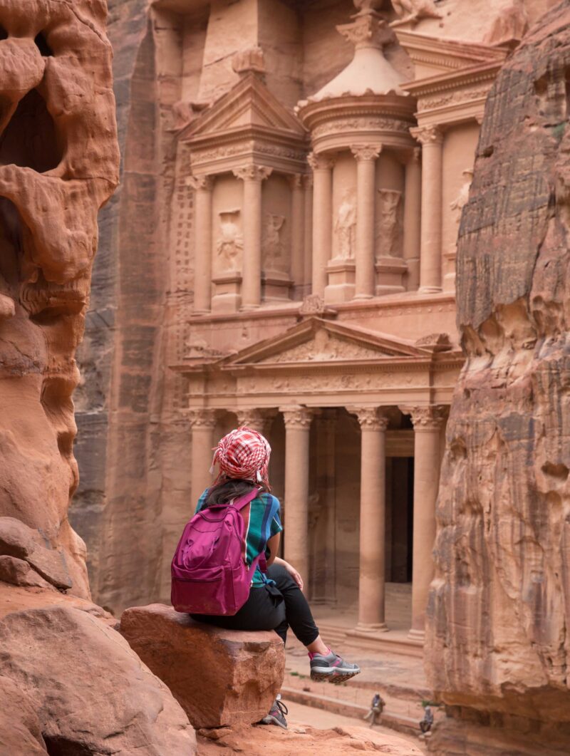 A traveller sits on a rocky ledge looking out towards the carved façade of Petra's Treasury in Jordan.