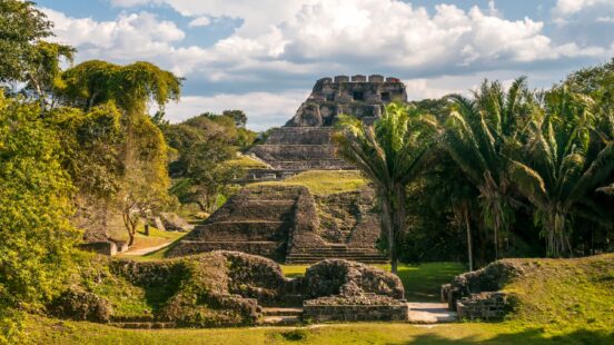 Large stone Mayan temple ruins surrounded by green trees and grass under a blue sky with clouds.