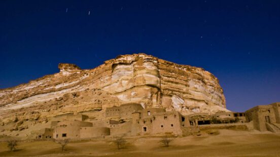 Mud-brick buildings at the foot of a massive, flat-topped desert mesa under a dark blue starry sky.