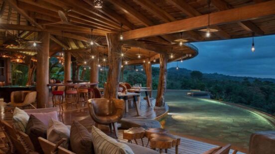 Upscale outdoor wooden terrace with hanging bulb lights, comfortable chairs, and a view of distant forested hills.