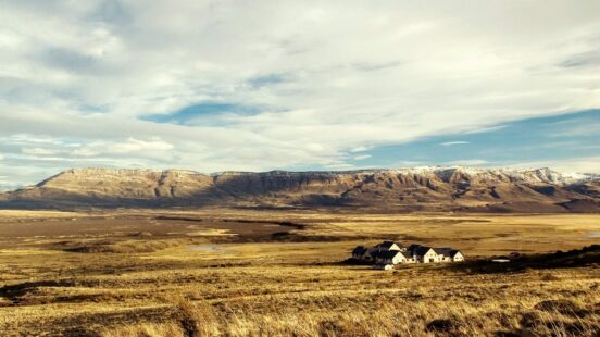 A group of white houses in a wide, grassy field with a large mountain range spanning the background.
