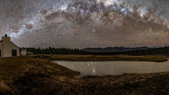 Panoramic view of the Milky Way galaxy over a dark landscape featuring a white cottage and a reflective pond.