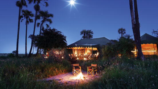 Night scene of a lighted safari tent and two chairs by a small campfire under a full moon and palm trees.