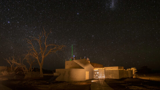 Small building at night under a vast starry sky with a green laser beam pointing upwards and a silhouette of a tree.