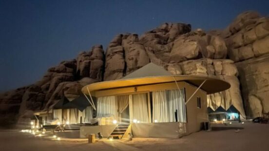 Illuminated luxury desert tents with pointed roofs situated in front of a large sandstone rock wall at dusk.