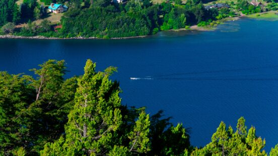 High-angle view of a vibrant blue lake with a small boat wake, bordered by green trees and distant houses.