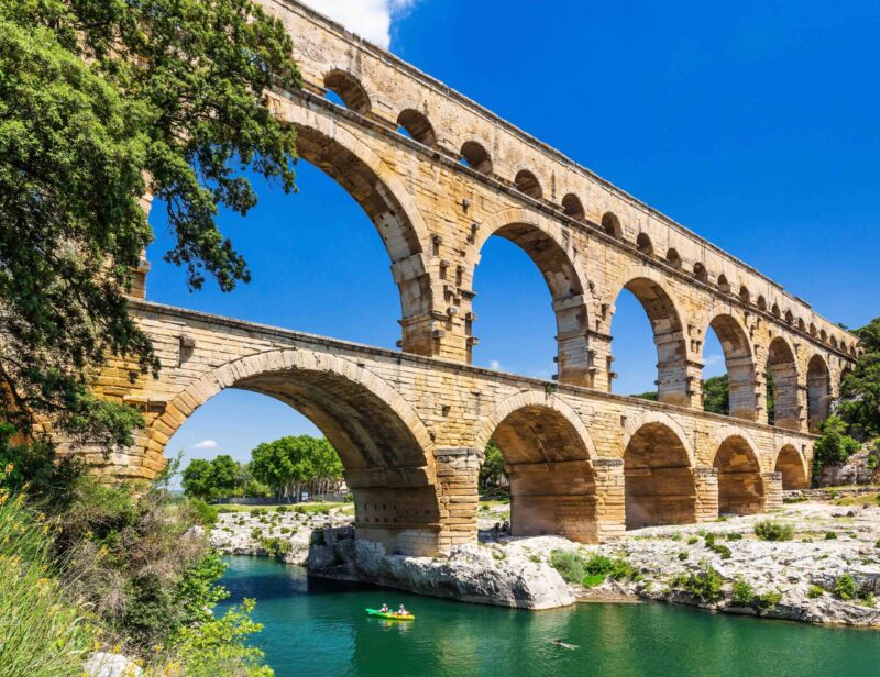 A high-angle view of the three-tiered Pont du Gard Roman aqueduct crossing a river with a green kayak below.