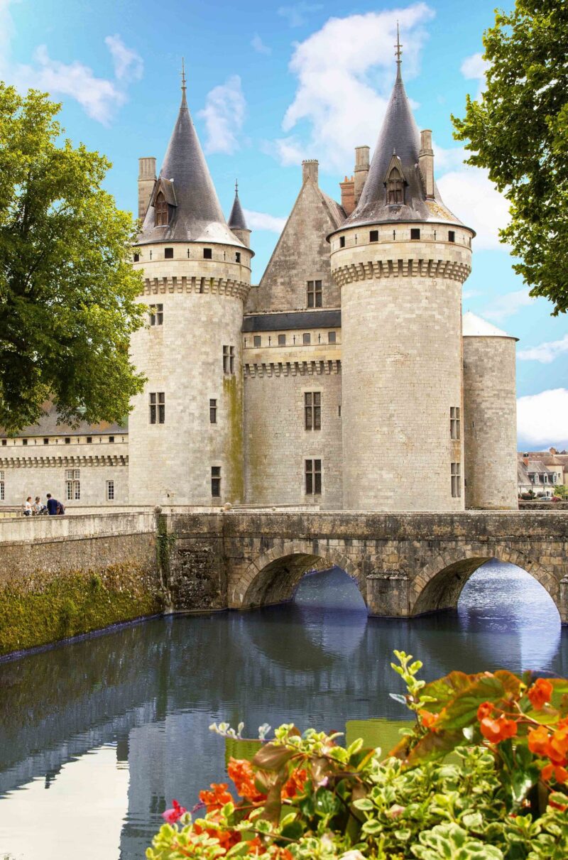 A French medieval castle with grey stone towers and a moat, viewed behind a stone bridge and colorful flowers.