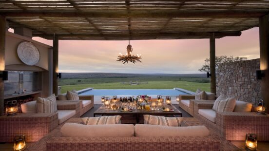 Luxury outdoor patio with wicker seating, lanterns, and an infinity pool overlooking a field at sunset.