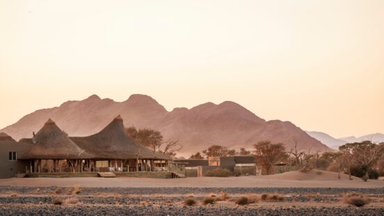 A luxury safari lodge with conical thatched roofs in a desert landscape at the base of large mountains.