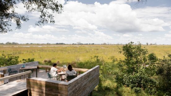 Two people sitting at a small table on a elevated wooden verandah looking over African bushland