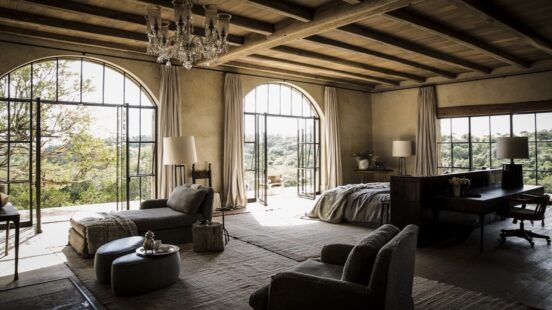 Interior of a high-end bedroom with large arched windows, a chandelier, and neutral-toned furniture.