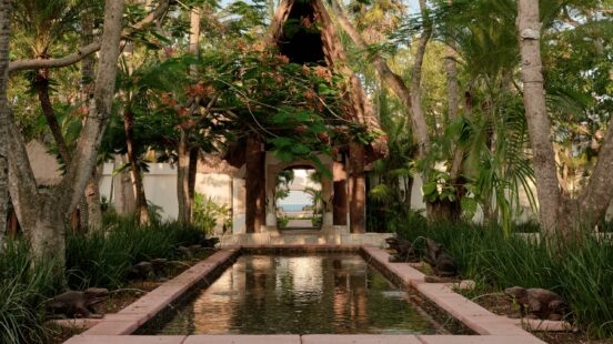 A symmetrical garden scene with a central water feature, frog statues, and a thatched pavilion framed by trees.