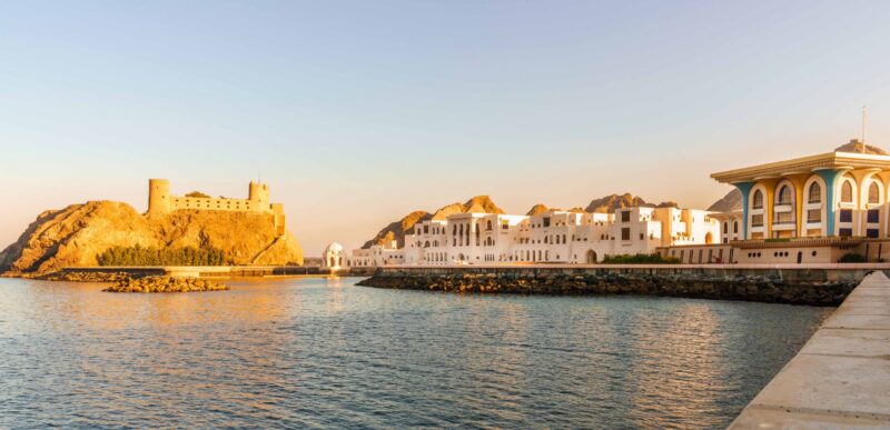 A scenic coastal view of a fort and white palace buildings at sunset on luxury Oman tours.