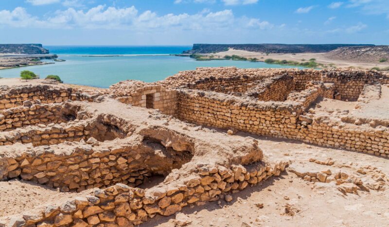 Stone ruins overlooking a blue coastal inlet and desert cliffs during luxury Oman trips.