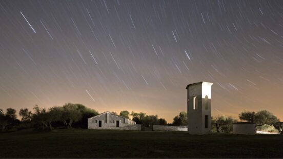 Diagonal white star trails across a night sky above a small white structure and a tall, thin tower in a dark field.