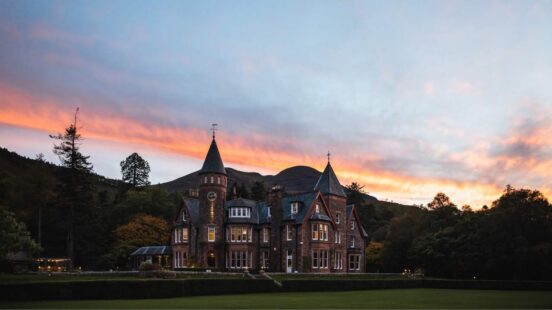 Large stone manor with a clock tower and pointed turrets set against a mountain range during a colorful sunset.