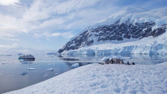 An expedition ship in a calm Antarctic bay surrounded by icebergs and high snowy peaks with penguins in the foreground.