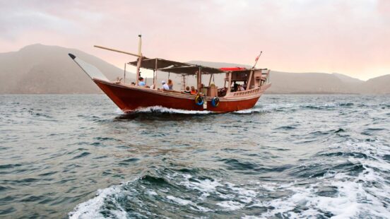 A wooden dhow boat with passengers sailing on the ocean with mountains visible in the misty background.