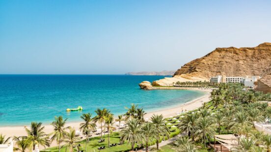 A sunny beach with turquoise water, palm trees, and a white hotel building nestled against rugged cliffs.
