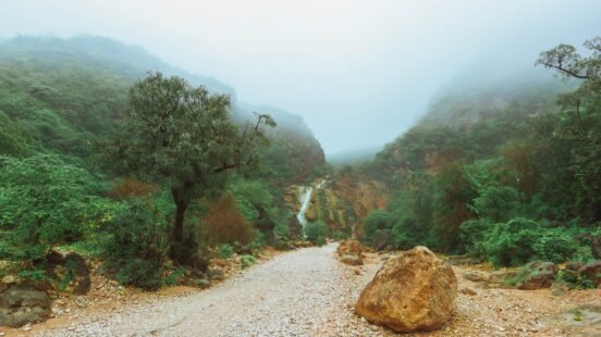 A rocky path in a green valley leading toward a waterfall under a thick foggy sky.