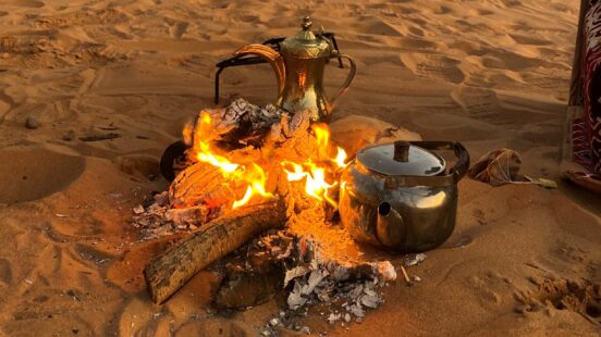 A golden coffee pot and silver kettle next to a small campfire on orange desert sand.