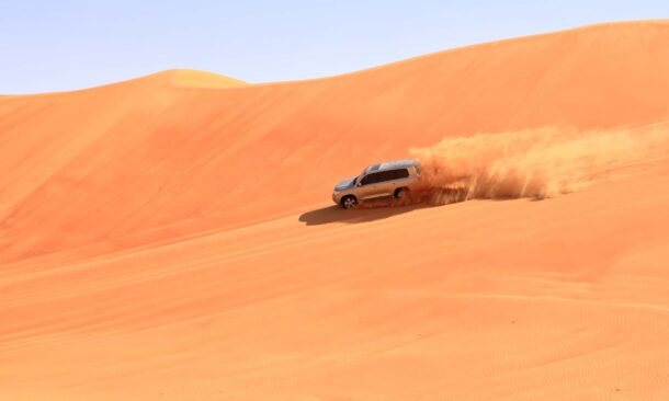 A silver vehicle maneuvers across a vast, rolling orange sand dune in a bright desert landscape.