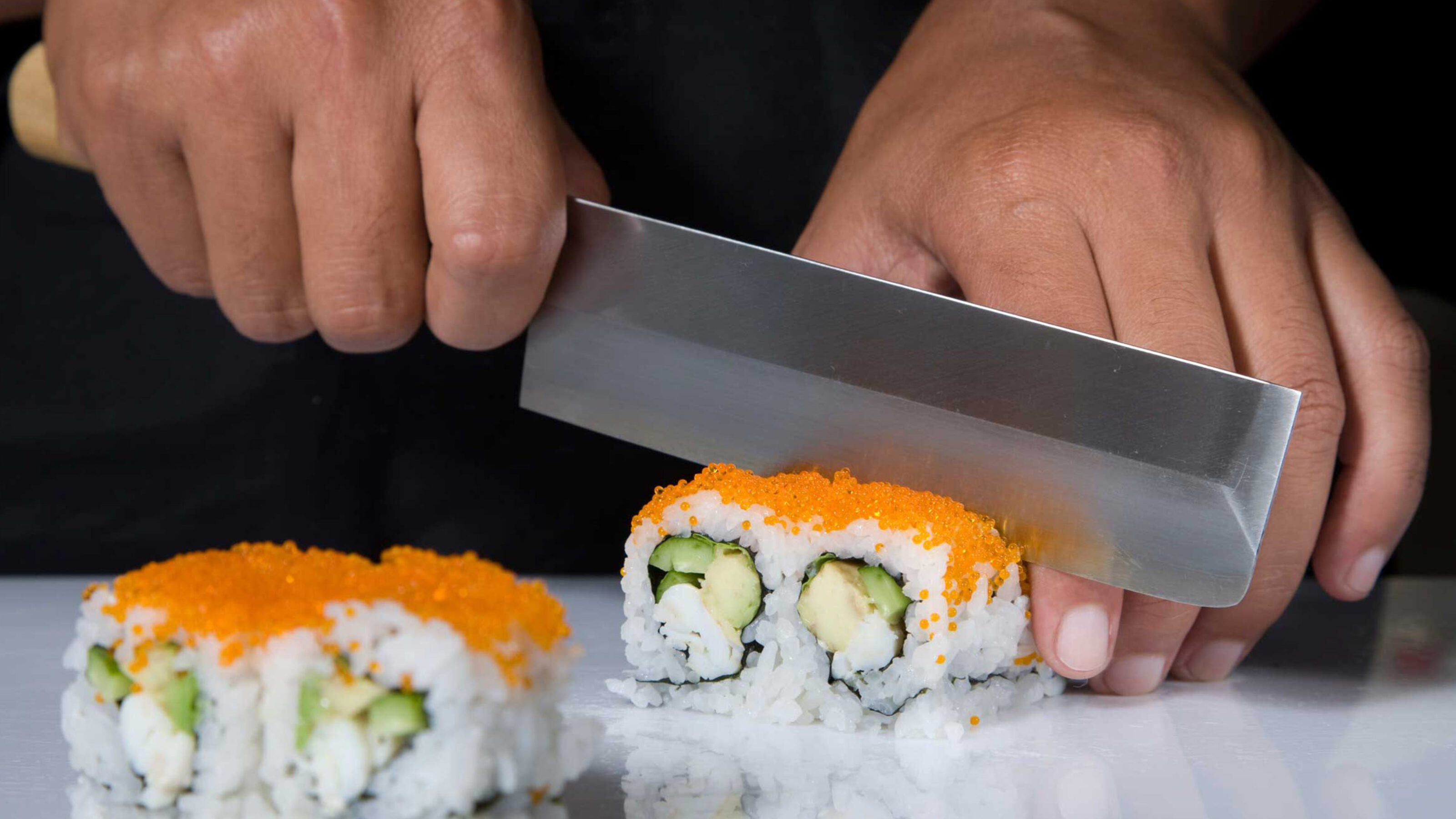 Close-up of a chef cutting sushi rolls covered in orange tobiko with a professional Japanese knife.
